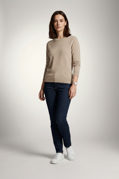 A woman stands against a light gray studio background wearing a beige crewneck sweater, dark blue slim-fit jeans, and white sneakers.