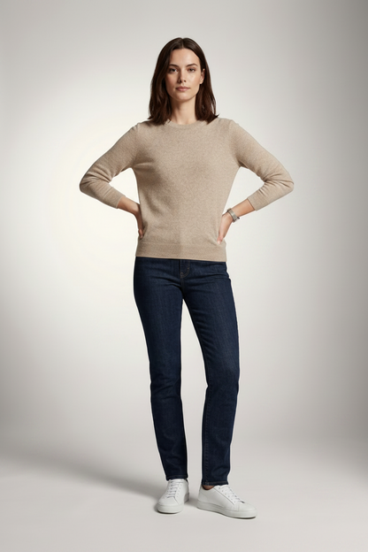 A woman stands against a light gray studio background wearing a beige crewneck sweater, dark blue slim-fit jeans, and white sneakers.