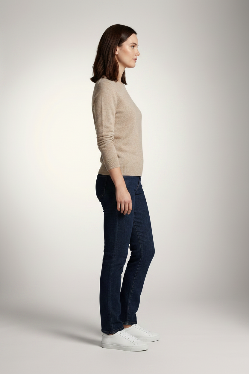 A woman stands against a light gray studio background wearing a beige crewneck sweater, dark blue slim-fit jeans, and white sneakers.