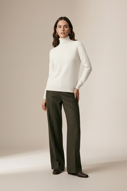 A woman stands with arms crossed wearing a cream ribbed turtleneck sweater and dark olive wide-leg trousers against a plain beige studio background.