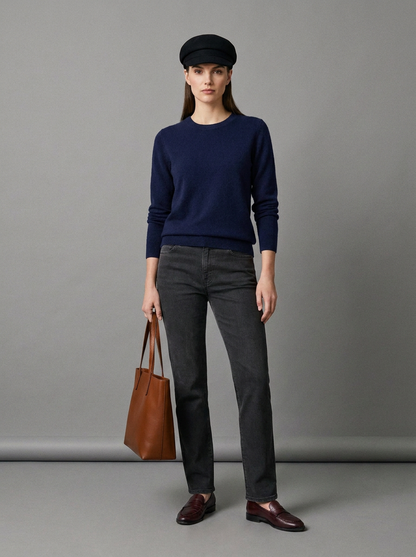 A woman stands against a plain gray studio background wearing a navy sweater, dark charcoal jeans, brown leather shoes, a black beret, and holding a brown leather tote bag.