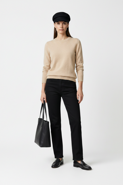 A model stands against a neutral gray studio background wearing a beige knit sweater, black trousers, a black beret, black loafers, and holding a black tote bag.