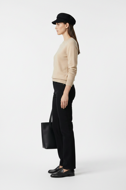 A model stands against a neutral gray studio background wearing a beige knit sweater, black trousers, a black beret, black loafers, and holding a black tote bag.