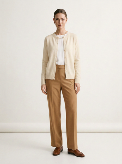 A woman stands against a plain off-white studio background wearing a cream cardigan over a white top, tan trousers, and brown loafers.
