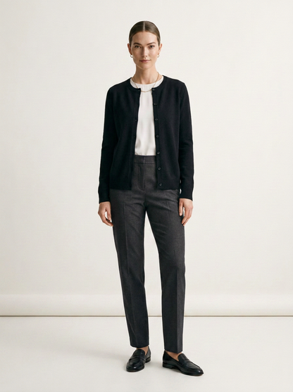 A woman stands in a studio wearing a black cardigan over a white top, dark gray trousers, and black loafers against a plain light background.