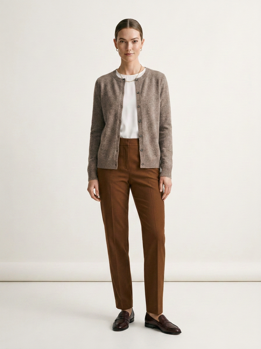A woman stands against a plain light beige studio background wearing a taupe knit cardigan over a white top, brown trousers, and dark brown loafers.