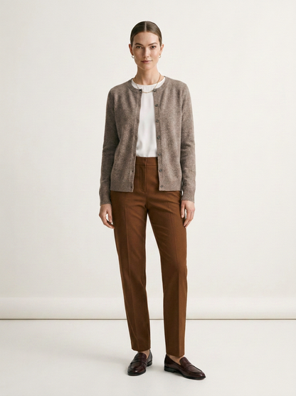 A woman stands against a plain light beige studio background wearing a taupe knit cardigan over a white top, brown trousers, and dark brown loafers.