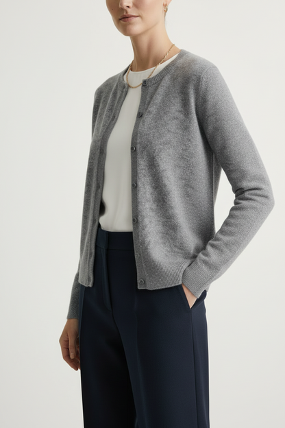 A close-up of a model wearing a heather gray knit cardigan with dark buttons over a white top and dark trousers, accessorized with a delicate gold chain necklace and hoop earrings.