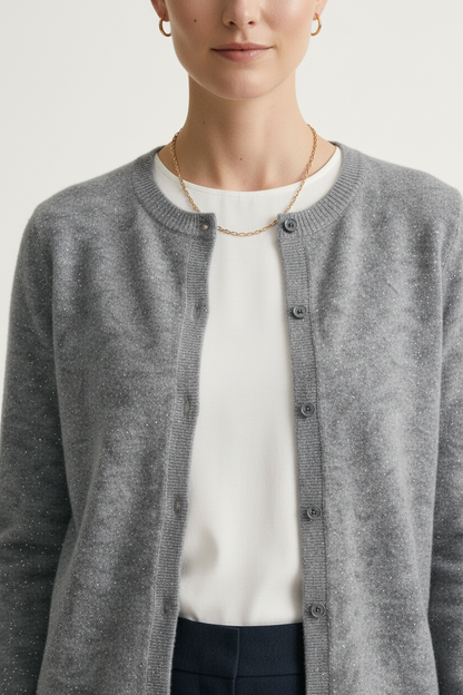 A close-up of a model wearing a heather gray knit cardigan with dark buttons over a white top and dark trousers, accessorized with a delicate gold chain necklace and hoop earrings.