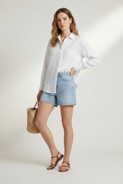 A woman stands against a light beige studio background wearing a white long-sleeve button-up shirt, light blue denim shorts, brown sandals, and holding a woven straw tote bag.