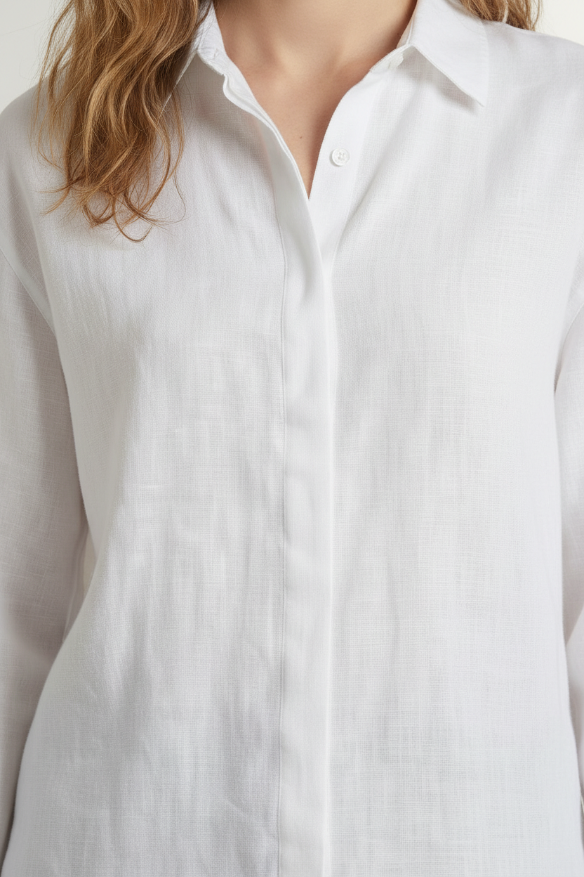 A woman stands against a light beige studio background wearing a white long-sleeve button-up shirt, light blue denim shorts, brown sandals, and holding a woven straw tote bag.