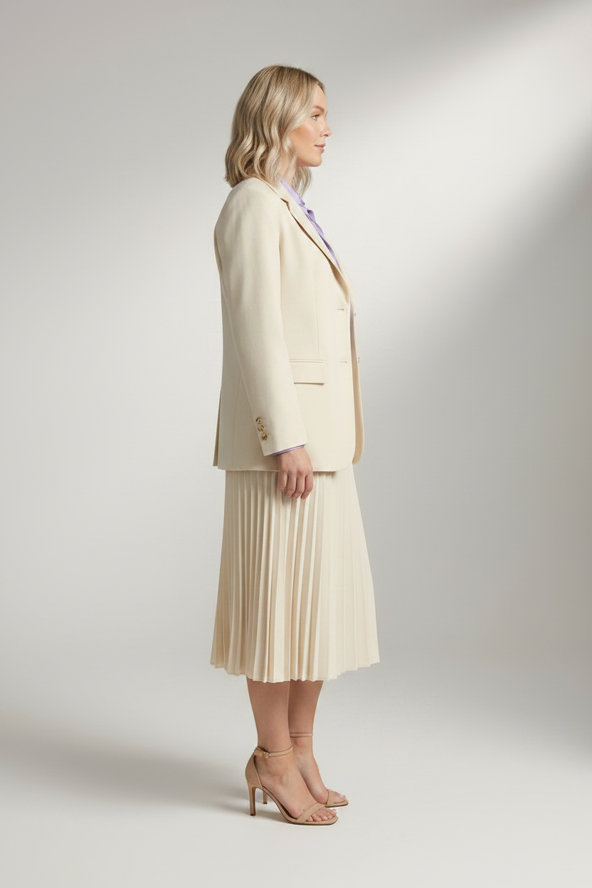A woman stands in a studio wearing a tailored cream blazer, matching pleated midi skirt, lavender button-up shirt, and nude high-heel sandals against a light gray backdrop.