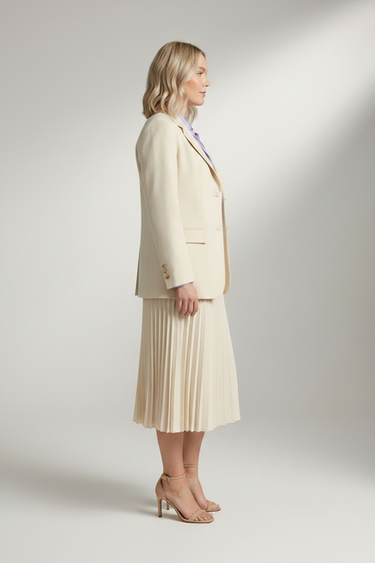 A woman stands in a studio wearing a tailored cream blazer, matching pleated midi skirt, lavender button-up shirt, and nude high-heel sandals against a light gray backdrop.