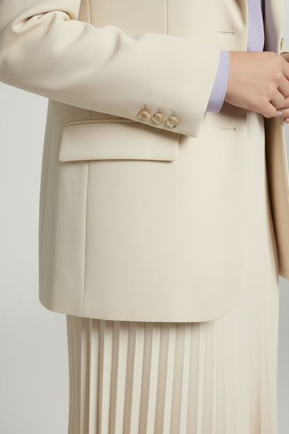 A woman models a matching cream-colored two-piece suit consisting of a tailored blazer and a pleated midi skirt, styled with a light purple patterned tie and beige high-heeled sandals against a neutral studio backdrop.
