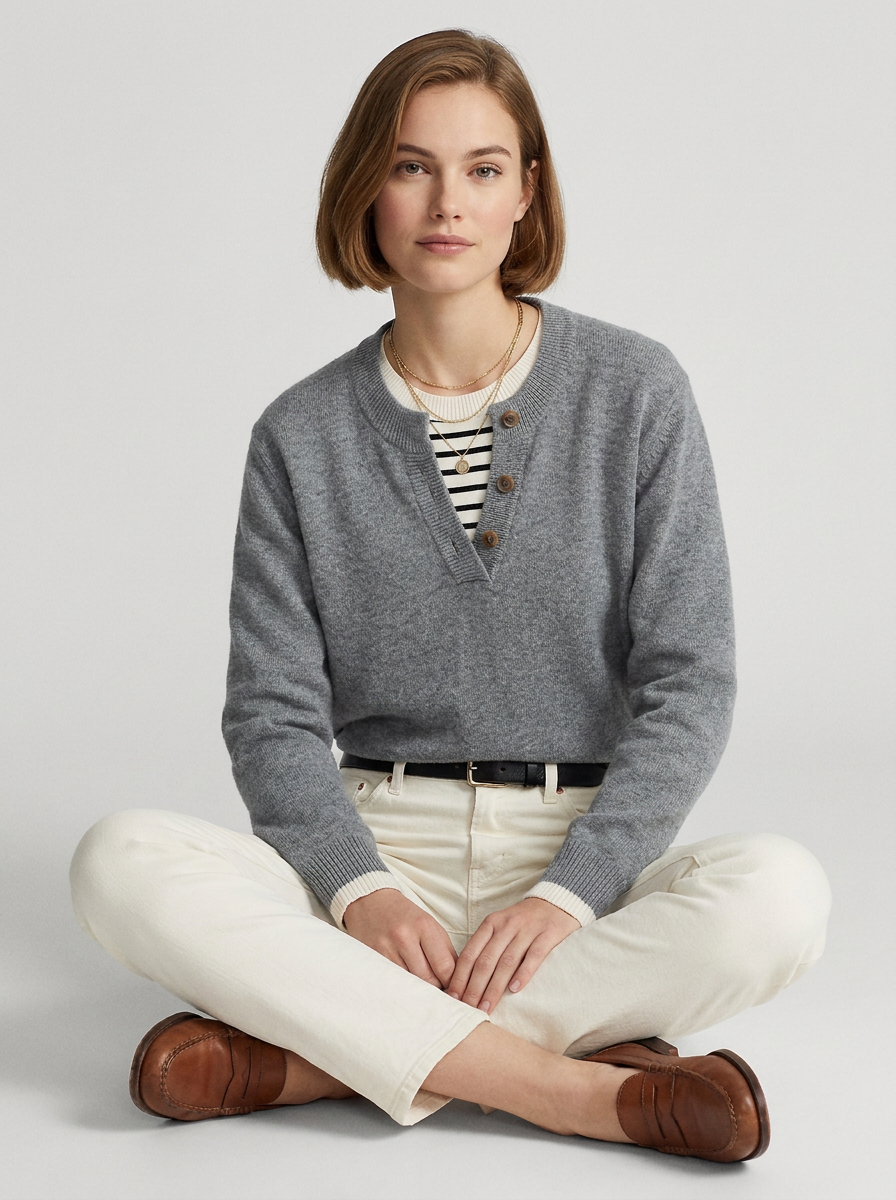A woman sits cross-legged on a light gray studio background wearing a heather gray V-neck knit sweater with brown buttons over a black-and-white striped shirt, cream pants, and brown leather shoes.