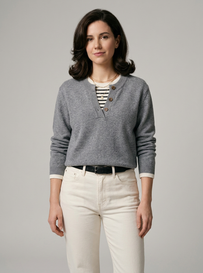 A woman modeling a gray long-sleeve henley sweater with a striped shirt and cream pants, photographed against a solid light gray studio background.