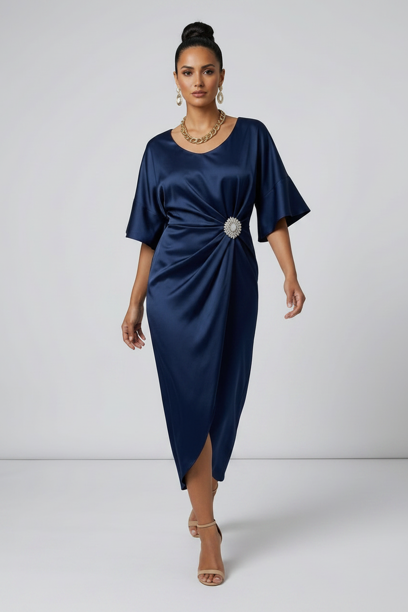 A woman models a navy blue satin midi dress with a gathered twist detail at the waist adorned with a jeweled brooch. The dress features short flutter sleeves and a high-low hemline, styled with nude high-heeled sandals against a light gray studio background.