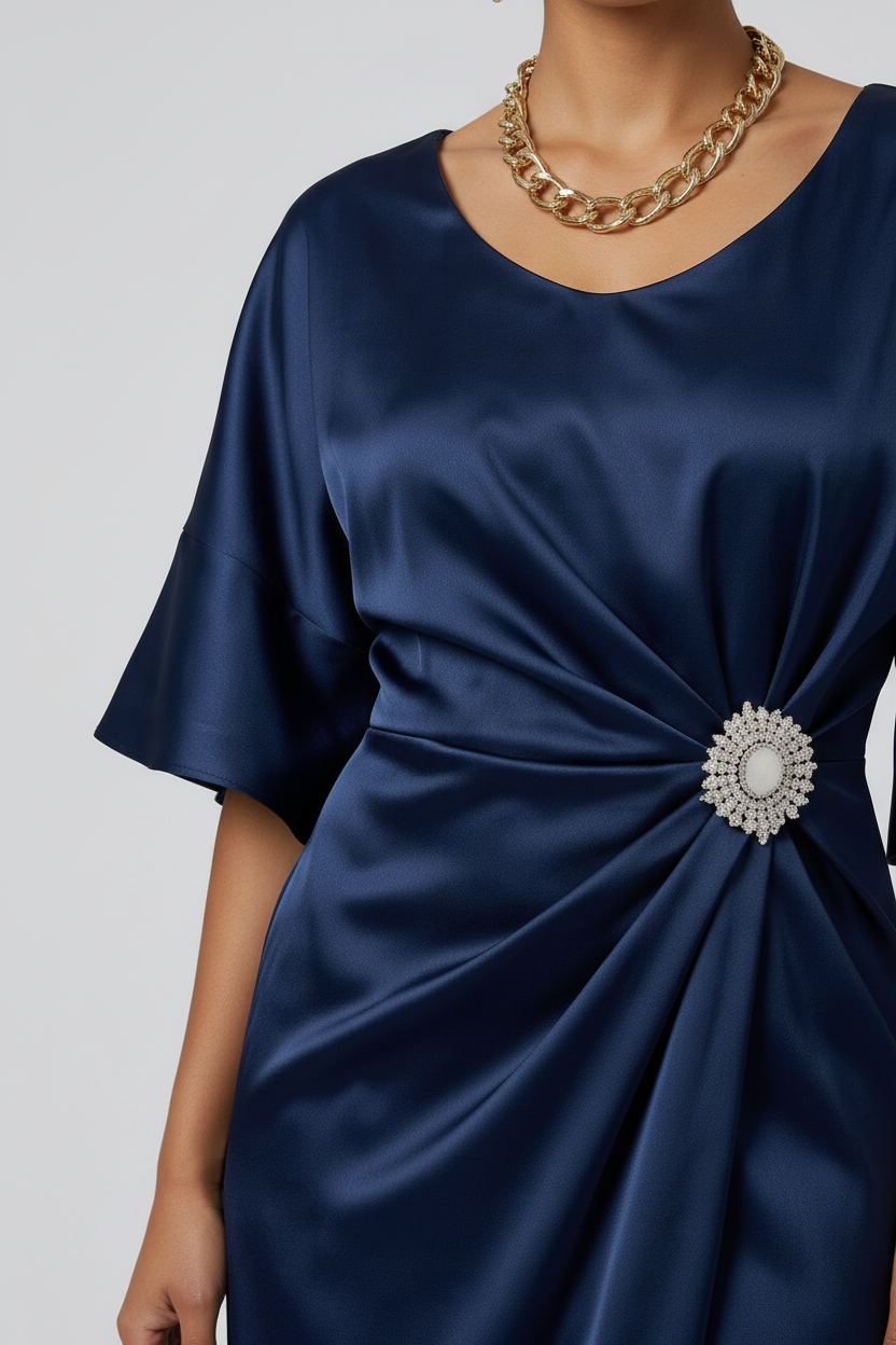 A woman models a navy blue satin midi dress with a gathered twist detail at the waist adorned with a jeweled brooch. The dress features short flutter sleeves and a high-low hemline, styled with nude high-heeled sandals against a light gray studio background.