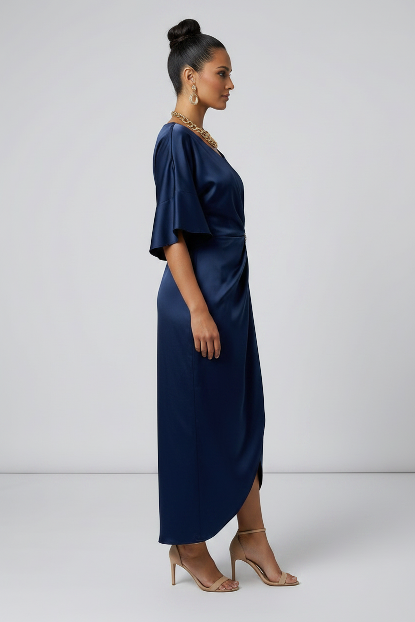 A woman models a navy blue satin midi dress with a gathered twist detail at the waist adorned with a jeweled brooch. The dress features short flutter sleeves and a high-low hemline, styled with nude high-heeled sandals against a light gray studio background.