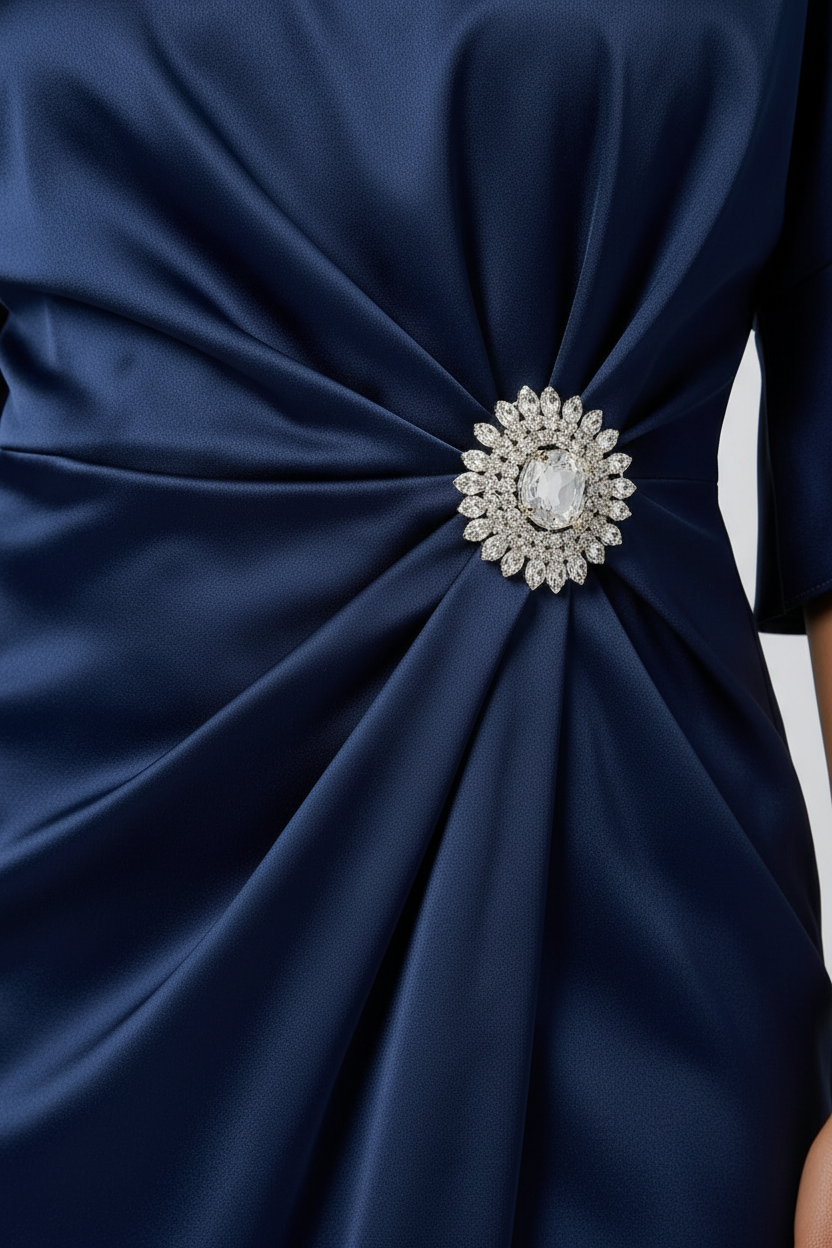 A woman models a navy blue satin midi dress with a gathered twist detail at the waist adorned with a jeweled brooch. The dress features short flutter sleeves and a high-low hemline, styled with nude high-heeled sandals against a light gray studio background.