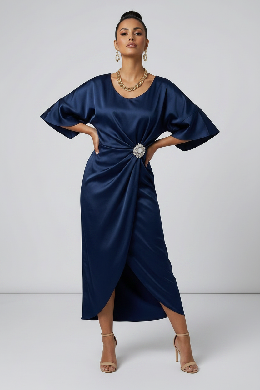 A woman models a navy blue satin midi dress with a gathered twist detail at the waist adorned with a jeweled brooch. The dress features short flutter sleeves and a high-low hemline, styled with nude high-heeled sandals against a light gray studio background.