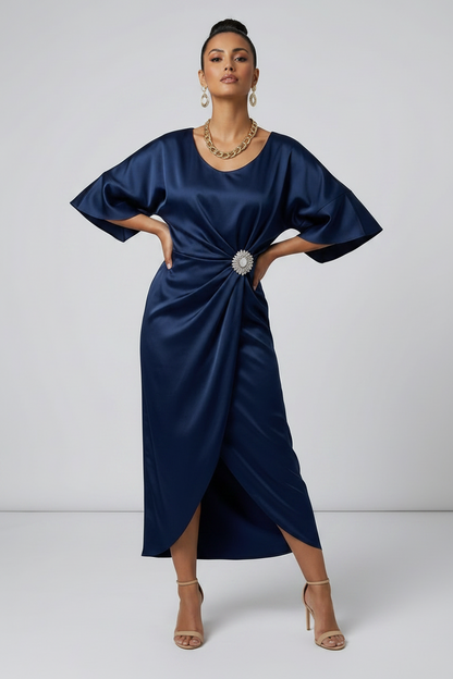 A woman models a navy blue satin midi dress with a gathered twist detail at the waist adorned with a jeweled brooch. The dress features short flutter sleeves and a high-low hemline, styled with nude high-heeled sandals against a light gray studio background.