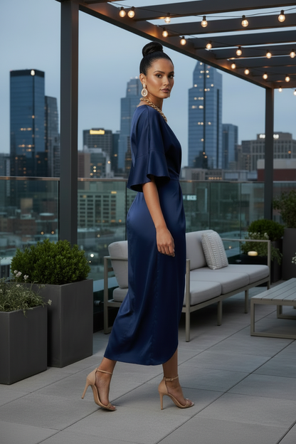 A woman stands on a rooftop patio at dusk wearing a navy blue satin midi dress with flutter sleeves and nude high heels. The background shows a city skyline, string lights, and outdoor furniture.