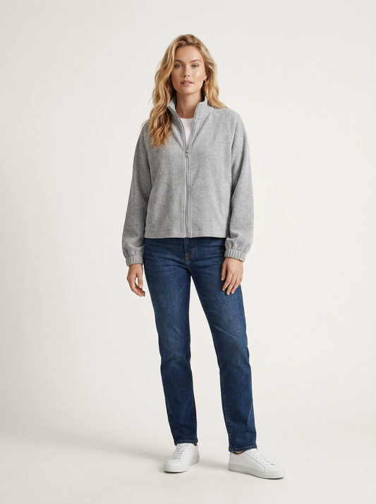 A woman modeling a light gray zip-up knit sweater, dark blue jeans, and white sneakers against a plain off-white studio background.