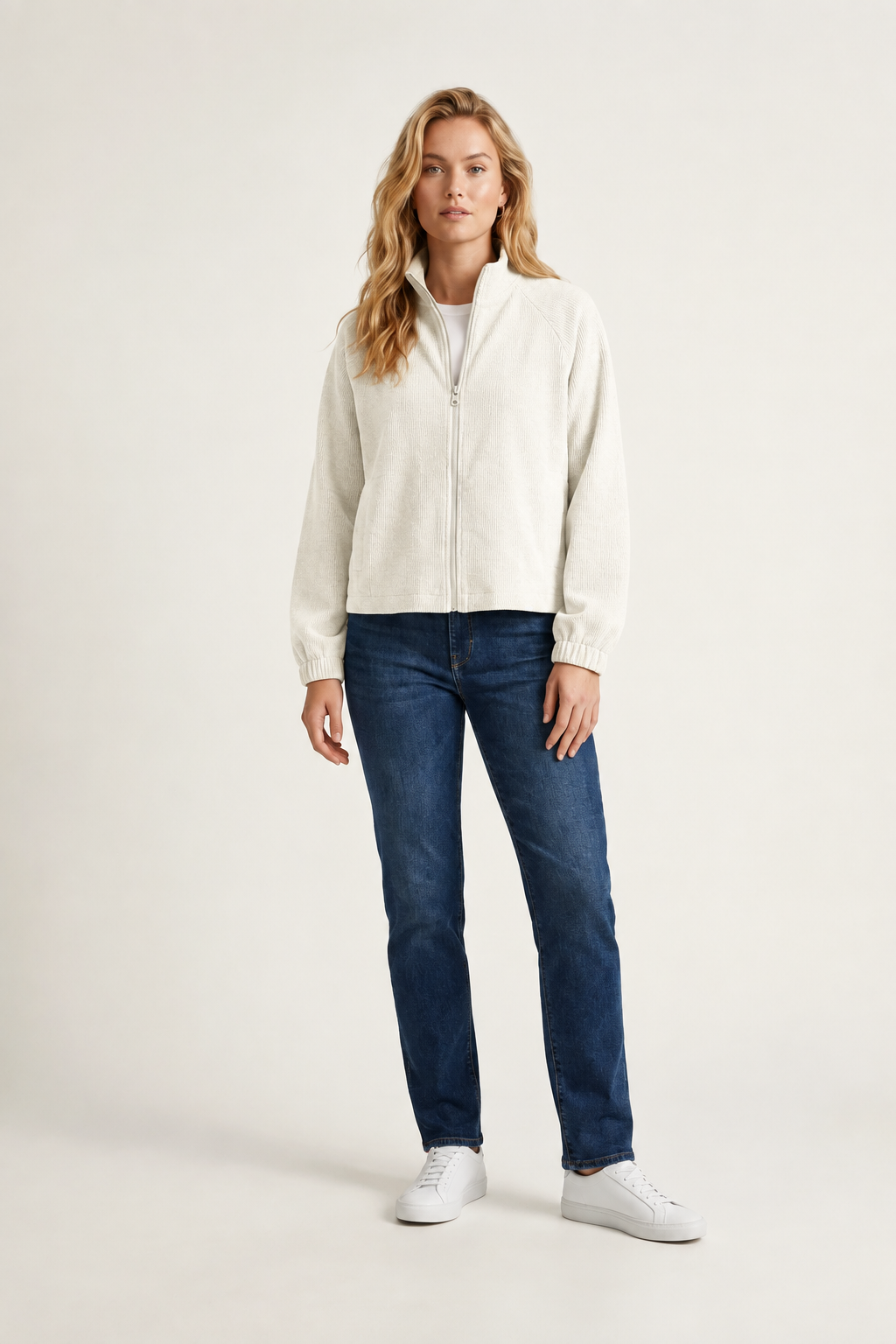 A woman modeling a light gray zip-up knit sweater, dark blue jeans, and white sneakers against a plain off-white studio background.