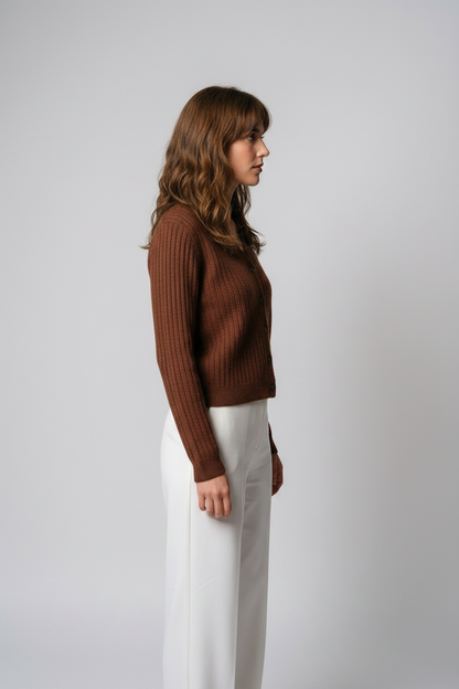 A woman stands against a light gray studio background wearing a brown ribbed button-front cardigan with a pointed collar and white wide-leg pants.