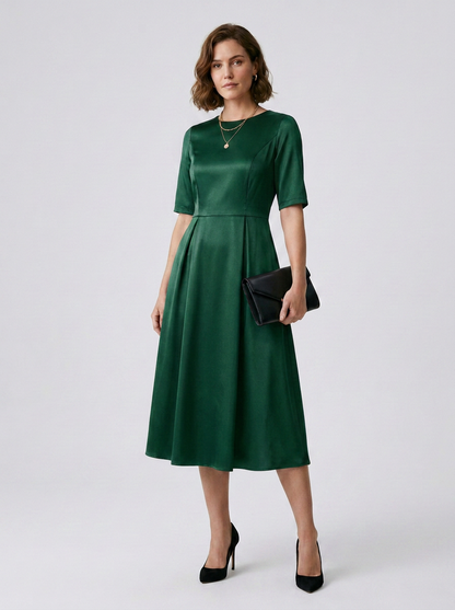 A woman models a dark green satin midi dress with short sleeves and a pleated skirt, holding a black clutch and wearing black heels against a light gray studio background.