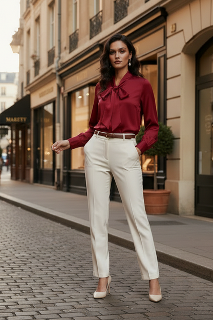 A woman stands on a cobblestone city street wearing a burgundy satin blouse with a bow collar and cream trousers, photographed in an urban outdoor setting.
