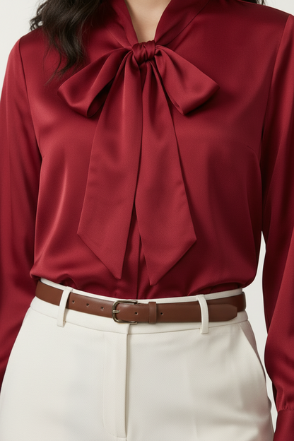 A woman modeling a long-sleeve satin-like blouse in deep burgundy with a large bow tie neckline, paired with off-white trousers and beige heels, against a plain light studio background.
