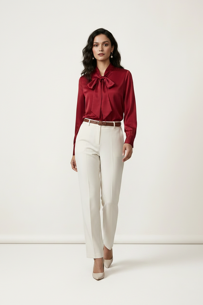 A woman modeling a long-sleeve satin-like blouse in deep burgundy with a large bow tie neckline, paired with off-white trousers and beige heels, against a plain light studio background.