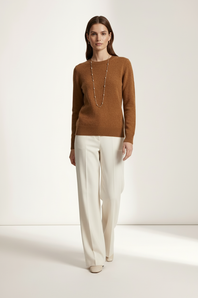 A woman modeling a warm brown crewneck knit sweater paired with off-white wide-leg trousers and a long beaded necklace, standing against a clean, light beige studio background.