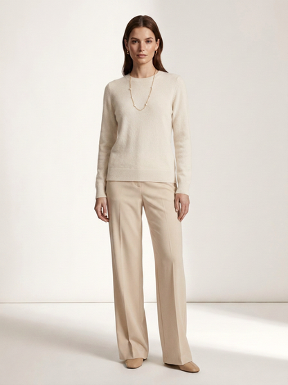 A woman stands in a studio wearing a cream knit long-sleeve top and matching wide-leg trousers, with a delicate gold necklace and beige flats.