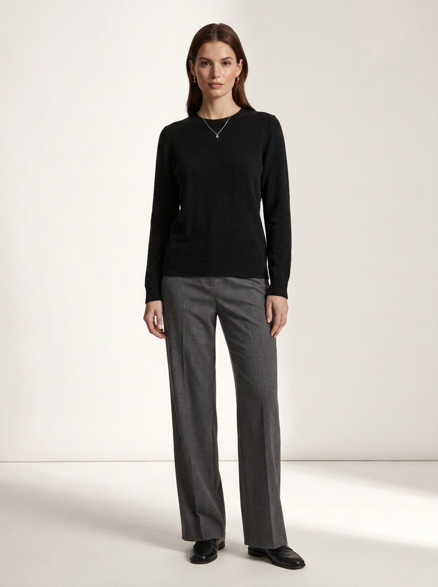 A woman stands against a plain light studio background wearing a black long-sleeve knit top, gray wide-leg trousers, and black shoes.