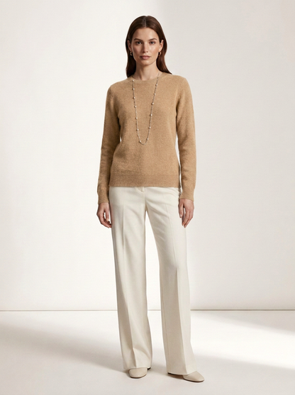 A woman stands in a studio wearing a beige knit sweater, off-white wide-leg trousers, and beige pointed shoes, with a long silver-toned necklace. The background is a clean warm off-white studio wall and floor.