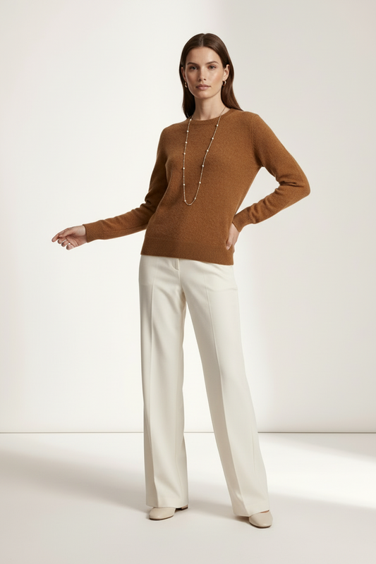 A woman modeling a long-sleeve brown knit sweater paired with off-white wide-leg trousers and a long beaded necklace, standing against a plain light beige studio background.