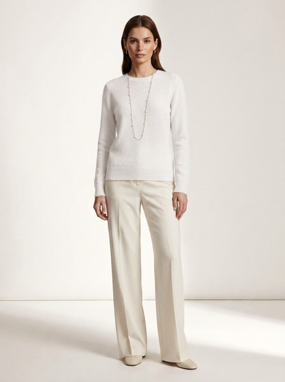 A woman stands in a studio wearing a white long-sleeve knit top, cream wide-leg trousers, and a long delicate necklace.