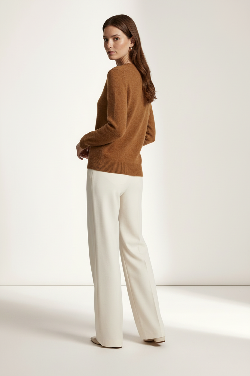 A woman stands in profile wearing a camel brown knit sweater and off-white wide-leg trousers against a clean, warm off-white studio background.
