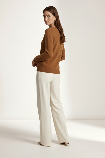A woman stands in profile wearing a camel brown knit sweater and off-white wide-leg trousers against a clean, warm off-white studio background.