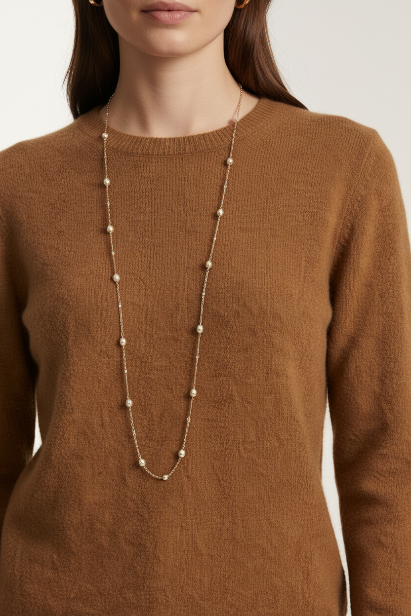 A close-up of a woman wearing a long, delicate necklace with small white pearls spaced along a thin gold-toned chain, paired with a brown knit sweater.