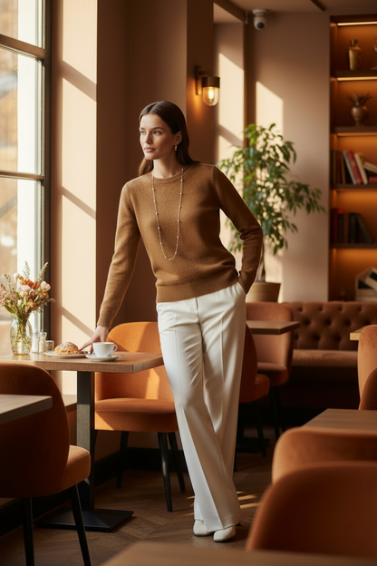 A woman stands in a warmly lit cafe wearing a brown knit sweater, cream wide-leg trousers, and white shoes, with a long silver necklace. Tables, chairs, shelves, and a window are visible in the background.