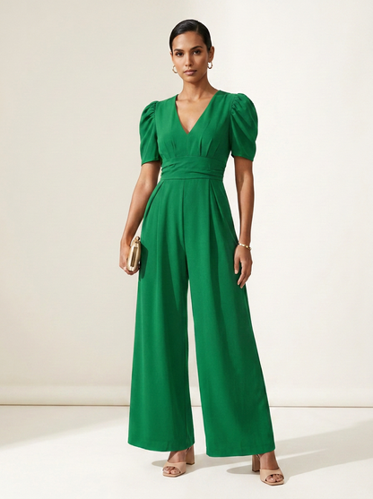 A woman modeling a bright green jumpsuit with puff sleeves, a V-neckline, and wide legs, standing against a plain off-white studio background.