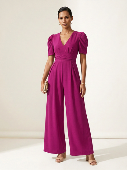 A woman modeling a bright magenta jumpsuit with puff sleeves, a wrap-style V-neck bodice, and wide legs, standing against a plain off-white studio background.