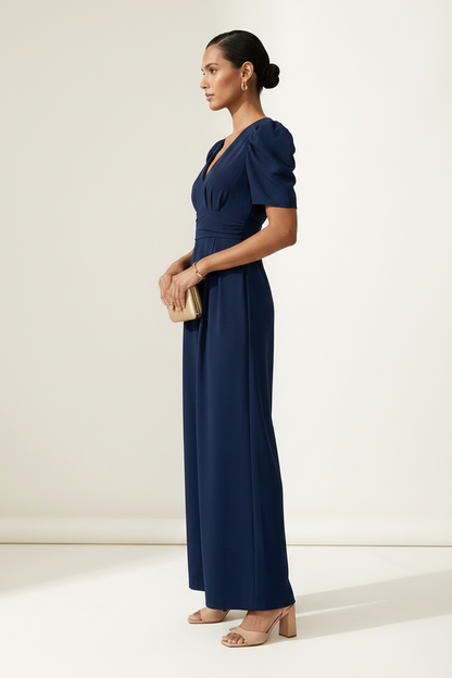 A woman modeling a navy blue jumpsuit with puff sleeves, a wrap-style V-neck bodice, and wide legs, standing against a plain off-white studio background.