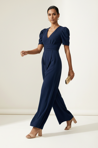 A woman modeling a navy blue jumpsuit with puff sleeves, a wrap-style V-neck bodice, and wide legs, standing against a plain off-white studio background.