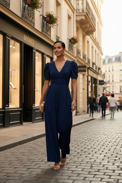 A woman modeling a navy blue jumpsuit with puff sleeves, a wrap-style V-neck bodice, and wide legs, standing against a plain off-white studio background.