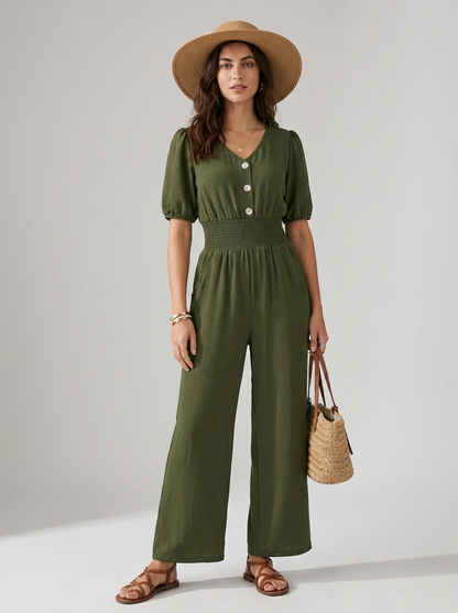 A woman models an olive green short-sleeve jumpsuit with puff sleeves, a buttoned V-neck, and wide legs. She is wearing a wide-brimmed tan hat, brown strappy sandals, and holding a woven straw handbag against a neutral studio background.